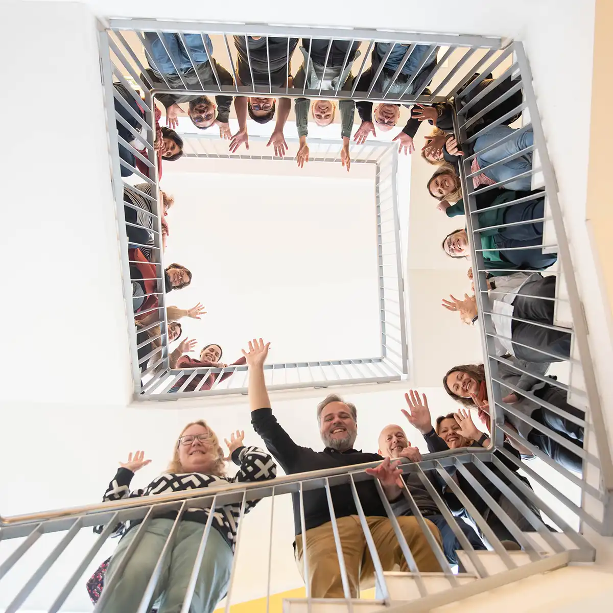 Menschen, die im Treppenhaus stehen, lachen und über das Treppengelände schauen und winken. Die Kamerapersepktive zeigt sie von unten