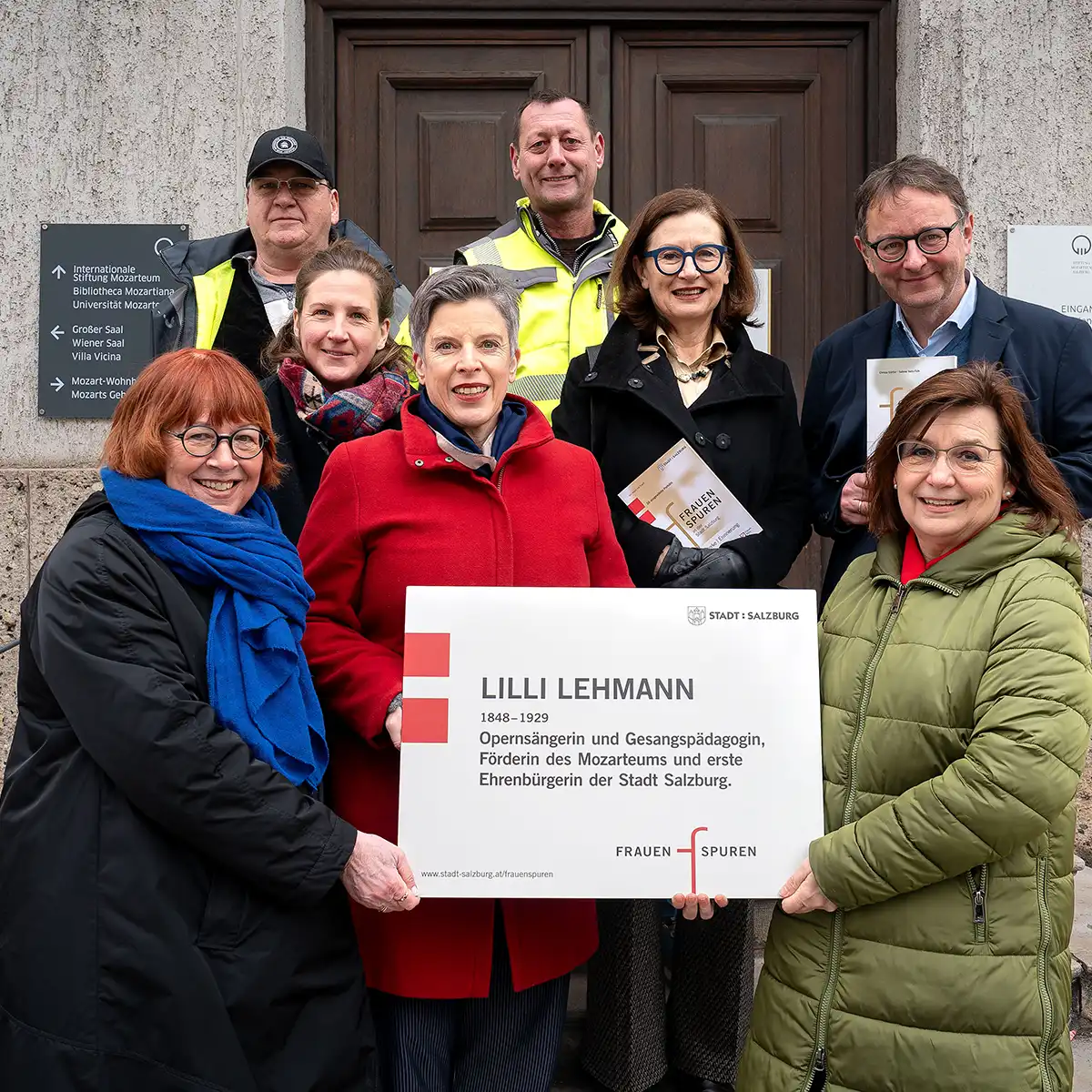 Eine Gruppe mittelalter Menschen steht vor einem Gebäude in Salzburg und hält einen Check oder eine Auszeichnung hoch. Sie lächeln