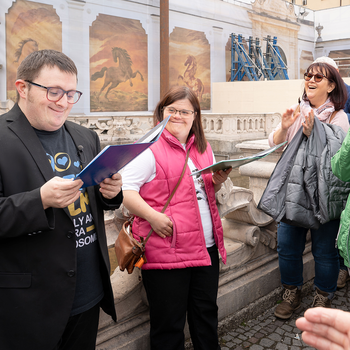 Ein Mann mit Behinderung lächelt und hält einen Vortrag vor Publikum. Die Leute freuen sich und klatschen in die Hände
