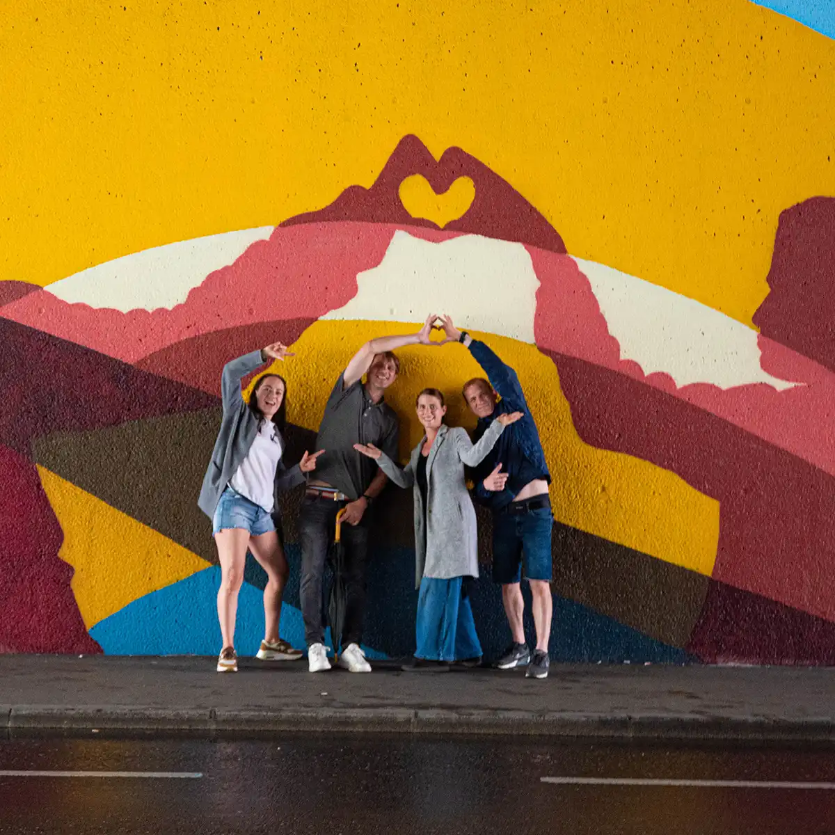 Vier Menschen stehen vor einer Grafittiwand und machen Herzen mit ihren Händen, sie lachen und freuen sich