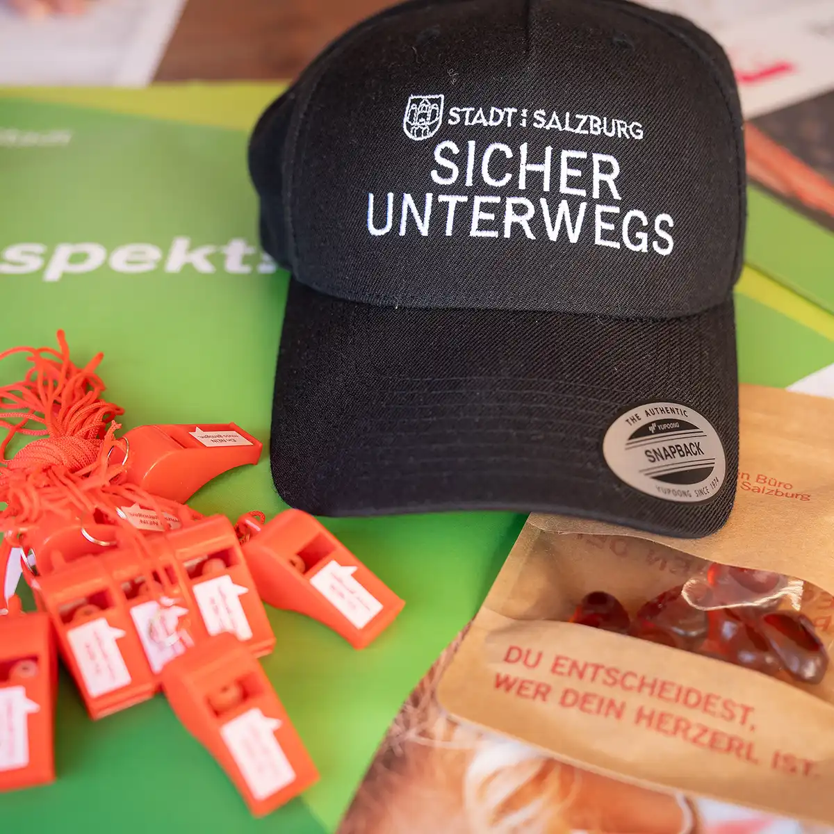 Ein schwarzes Capie mit der Aufschrift: sicher unterwegs, liegt neben roten Trillerpfeifen auf dem Tisch