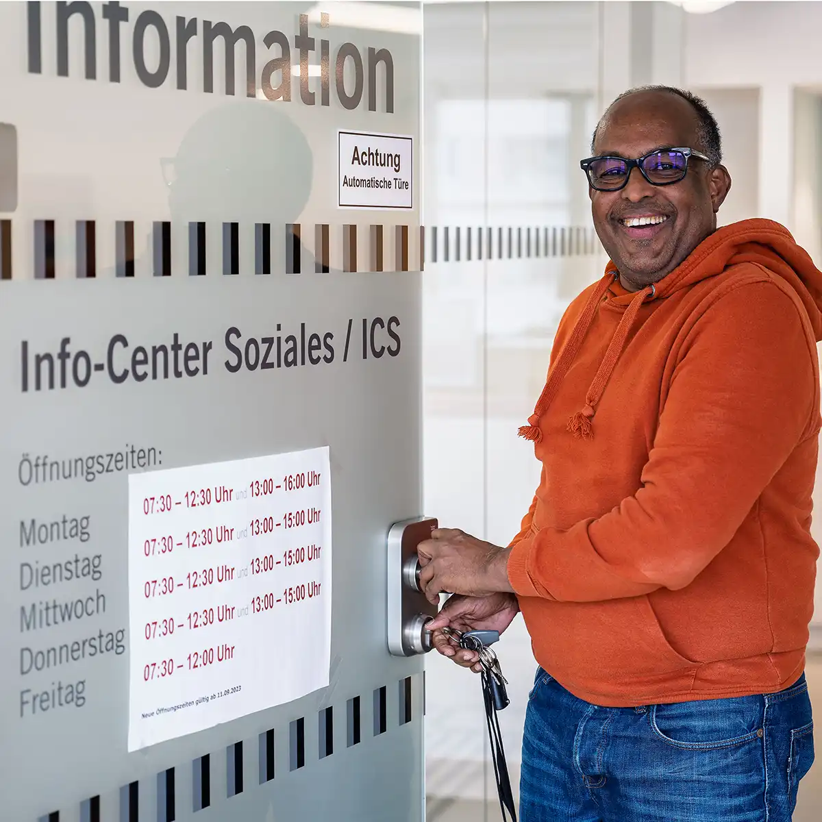 Ein Mann in orangener Jacke mit einem Lächeln im Gesicht schließt mit einem Schluss die Glastür vom Infocenter Soziales in Salzburg auf