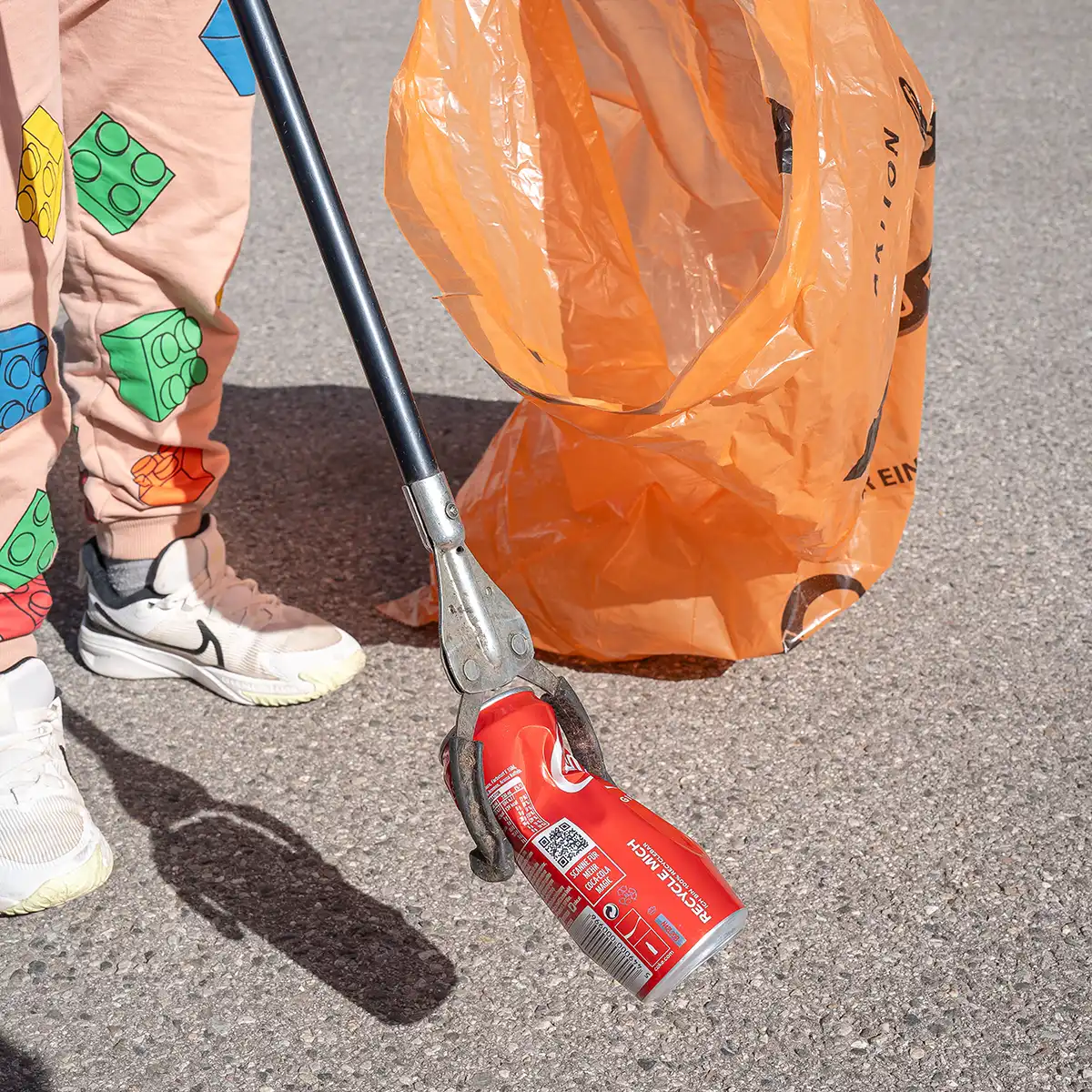 Eine Person hält einen Müllbeutel in der Hand und sammelt mit einem Stab eine Coladose vom Boden auf