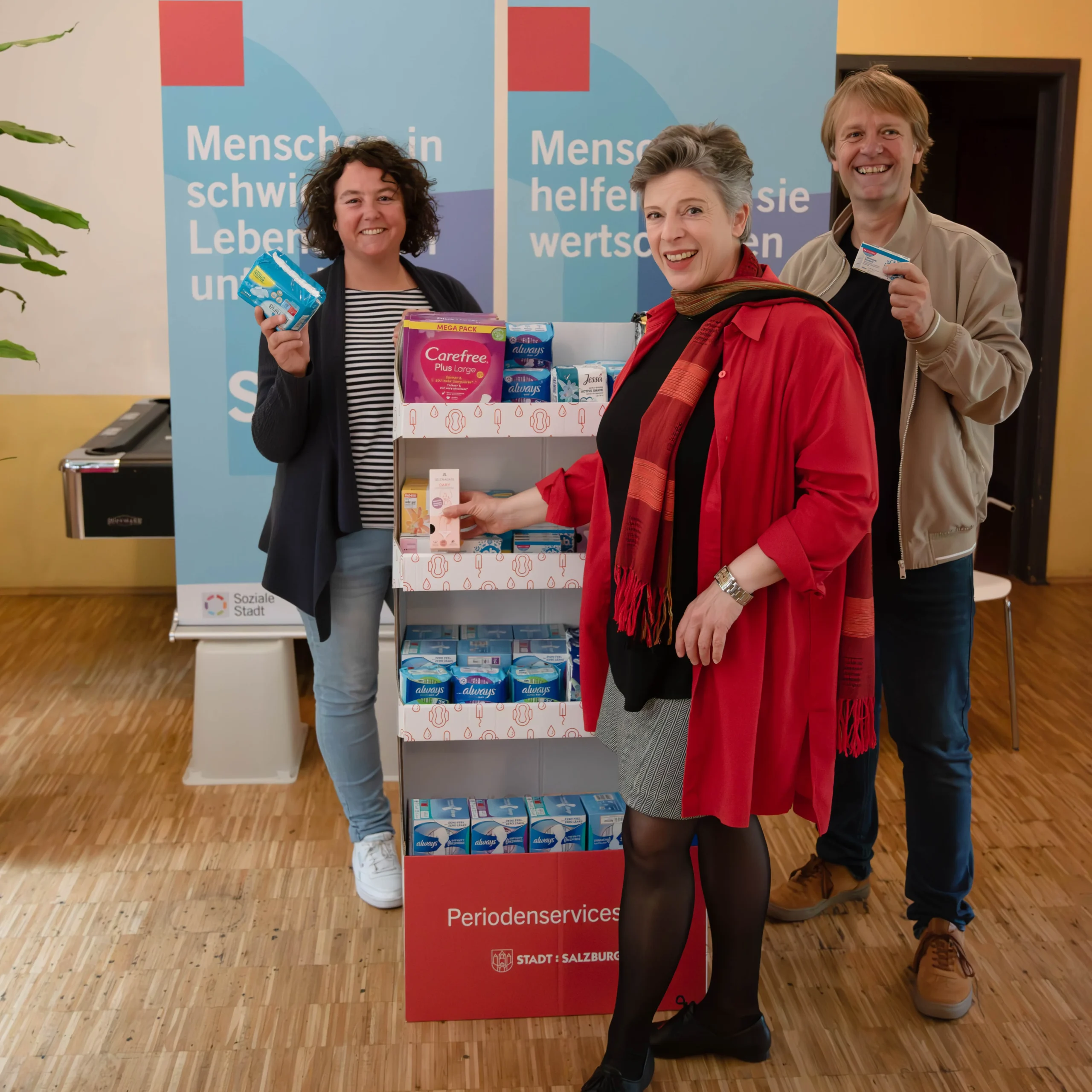 zwei Frauen und ein Mann stehen bei einem Aufsteller für Periodenprodukte