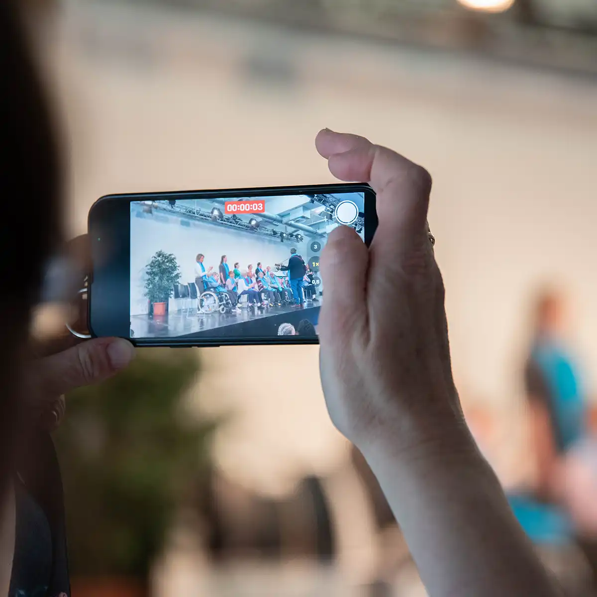 Eine Nahaufnahme von einer Hand, die ein Smartphone hält und filmt. Auf dem Handybildschirm sieht man Menschen im Rollstuhl