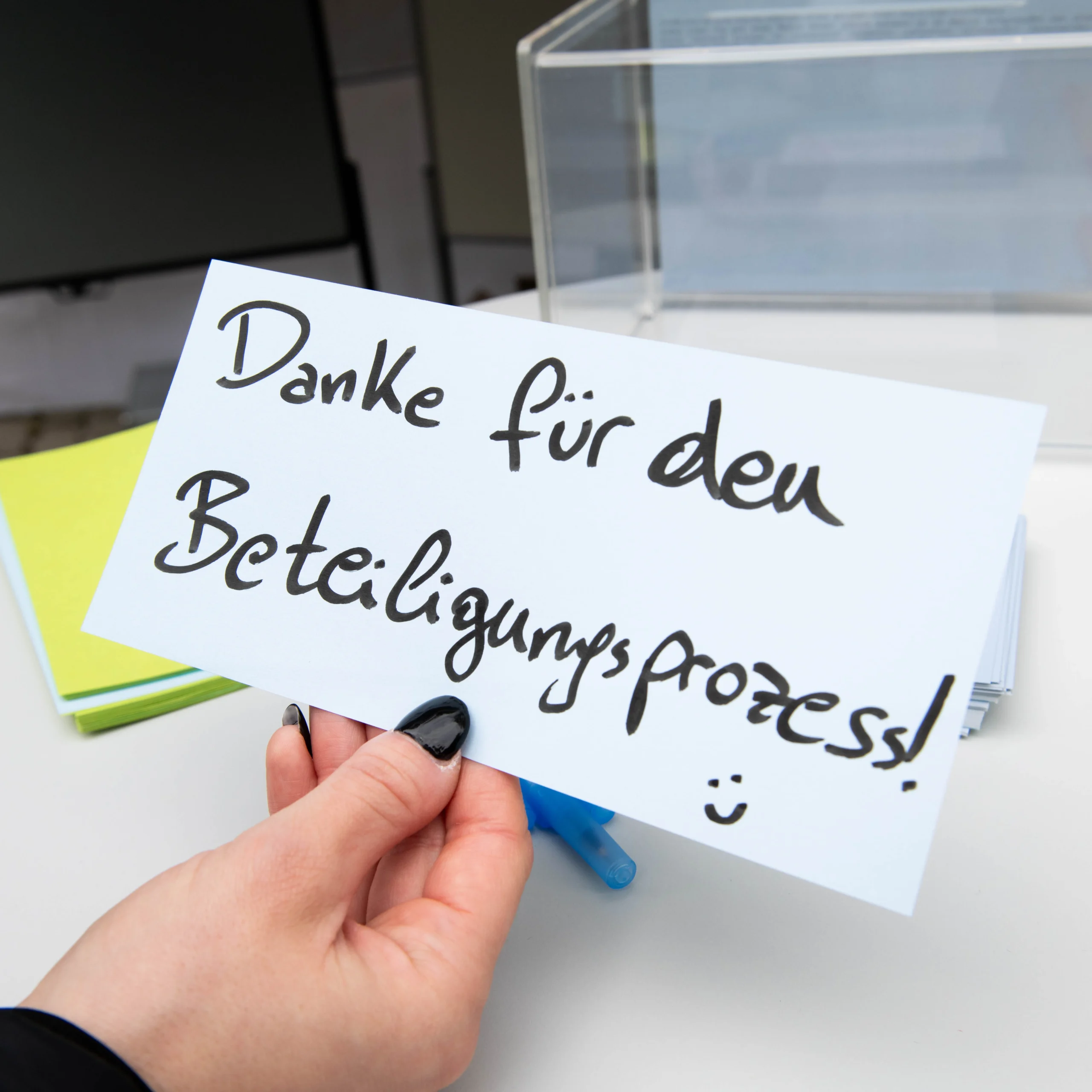 eine Dame mit schwarzen FIngernägeln hält einen Handgeschriebenen Zettel in der Hand mit der aufschrift: "Danke für den Beteiligungsprozess :)"