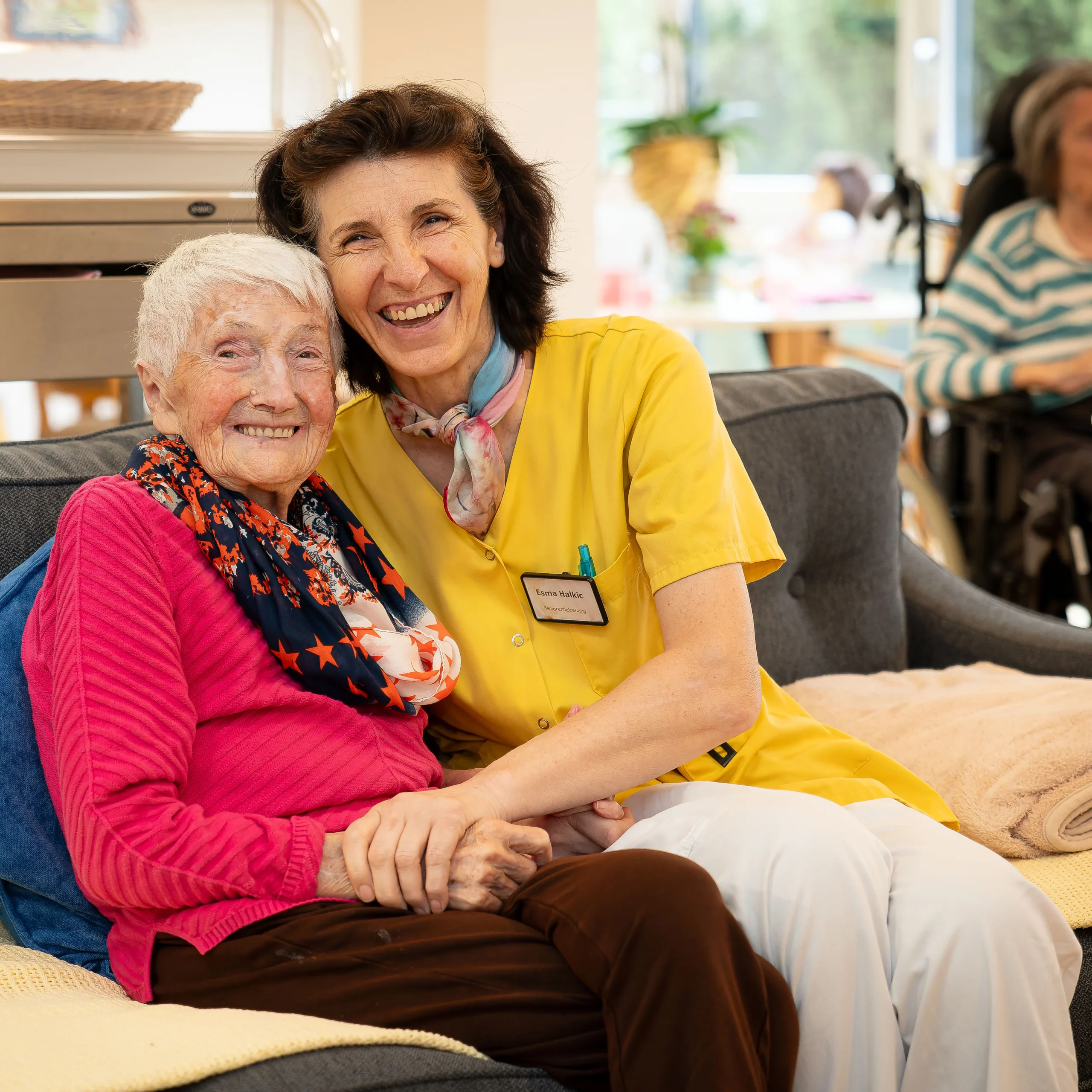 Eine Pflegerin aus dem Seniorenwohnheim und eine ältere Dame sitzen händchenhaltend auf einem Sofa und lachen