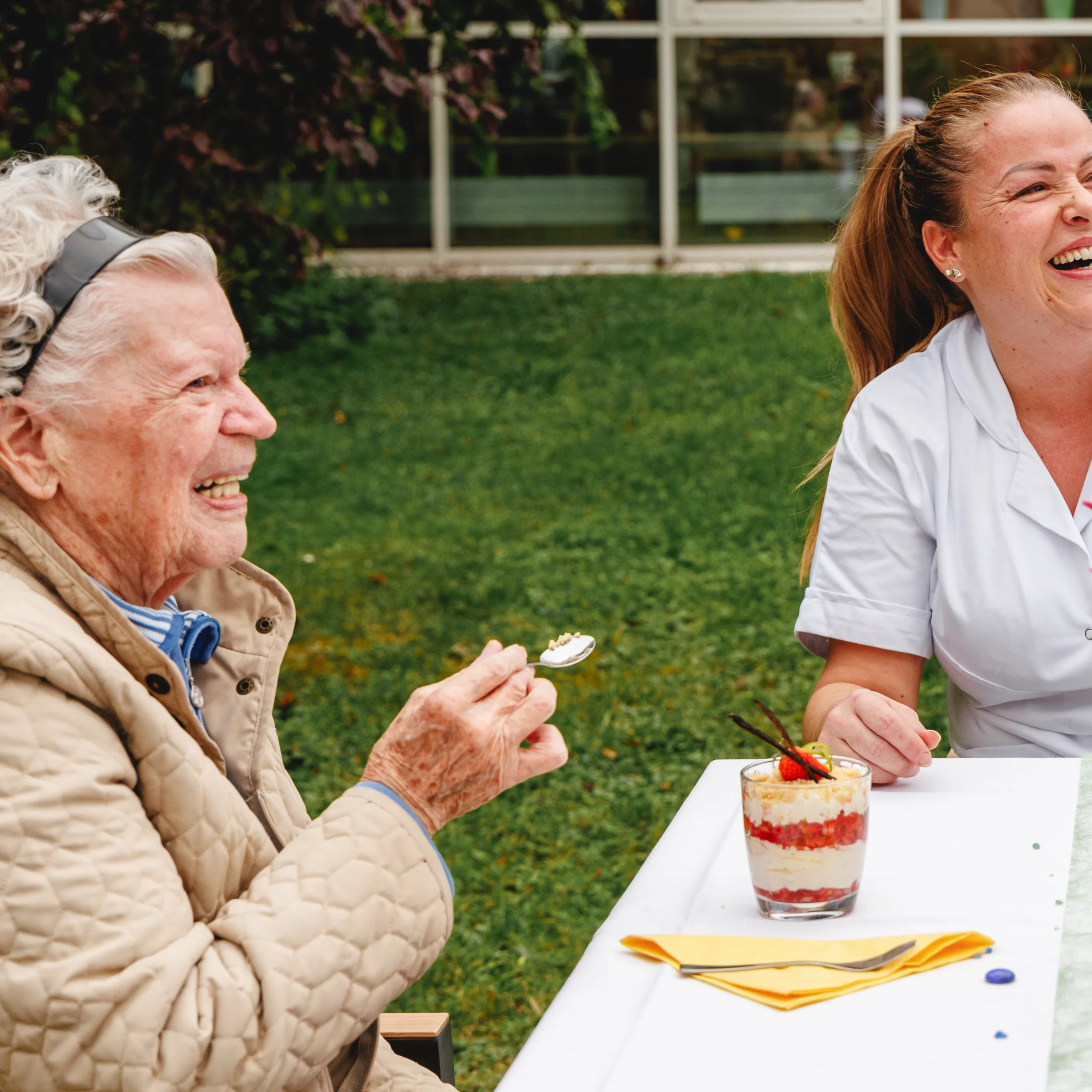 Eine alte Dame sitzt lachend mit einer Pflegerin des Seniorenwohnheimes an eineEine alte Dame sitzt lachend mit einer Pflegerin des Seniorenwohnheims an einem Tisch und genießt ein Dessert
