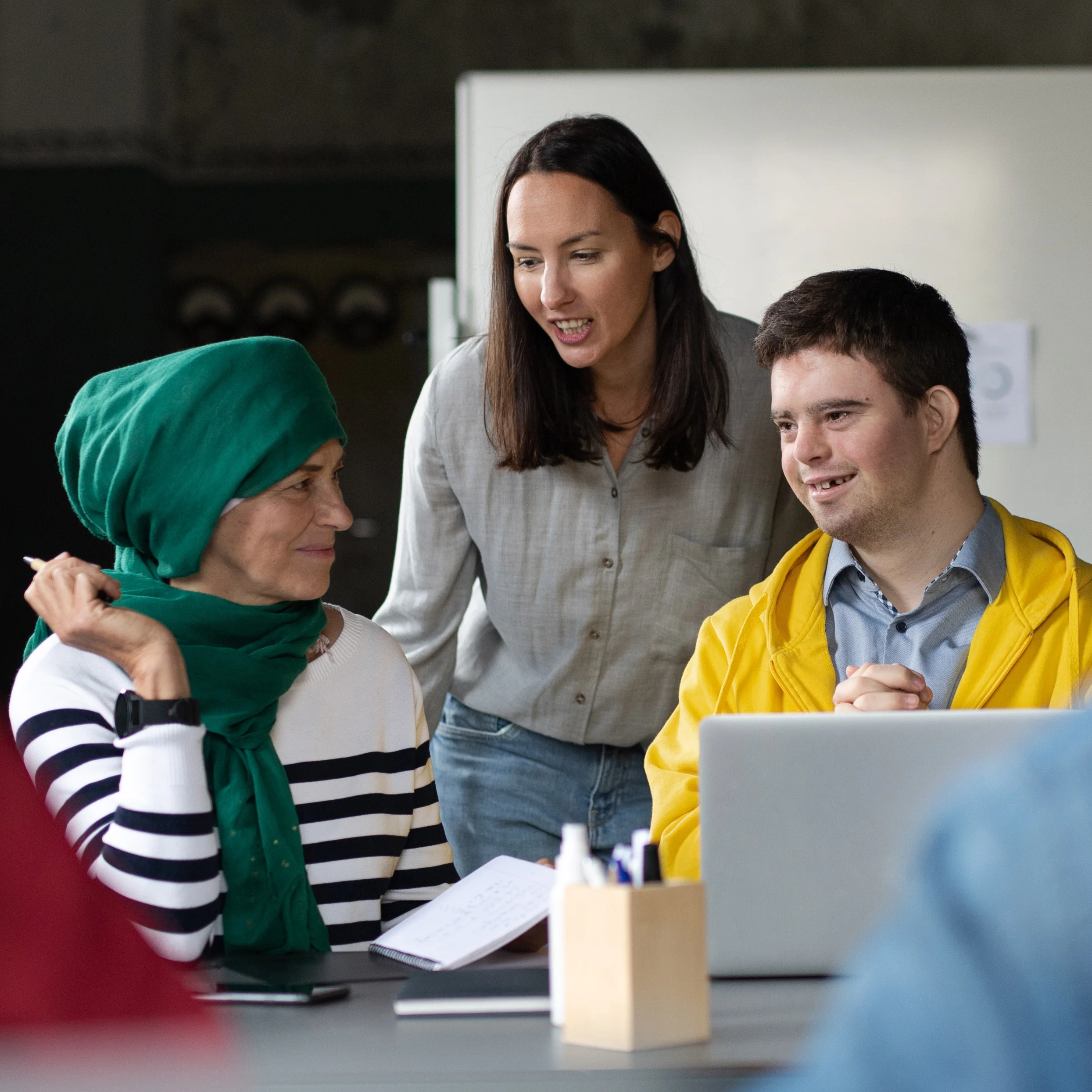 eine Frau mit Kopftuch und ein Junge mit Behinderung sitzen an einem Tisch, eine Sozialarbeiterin unterhält sich freundlich mit ihnen