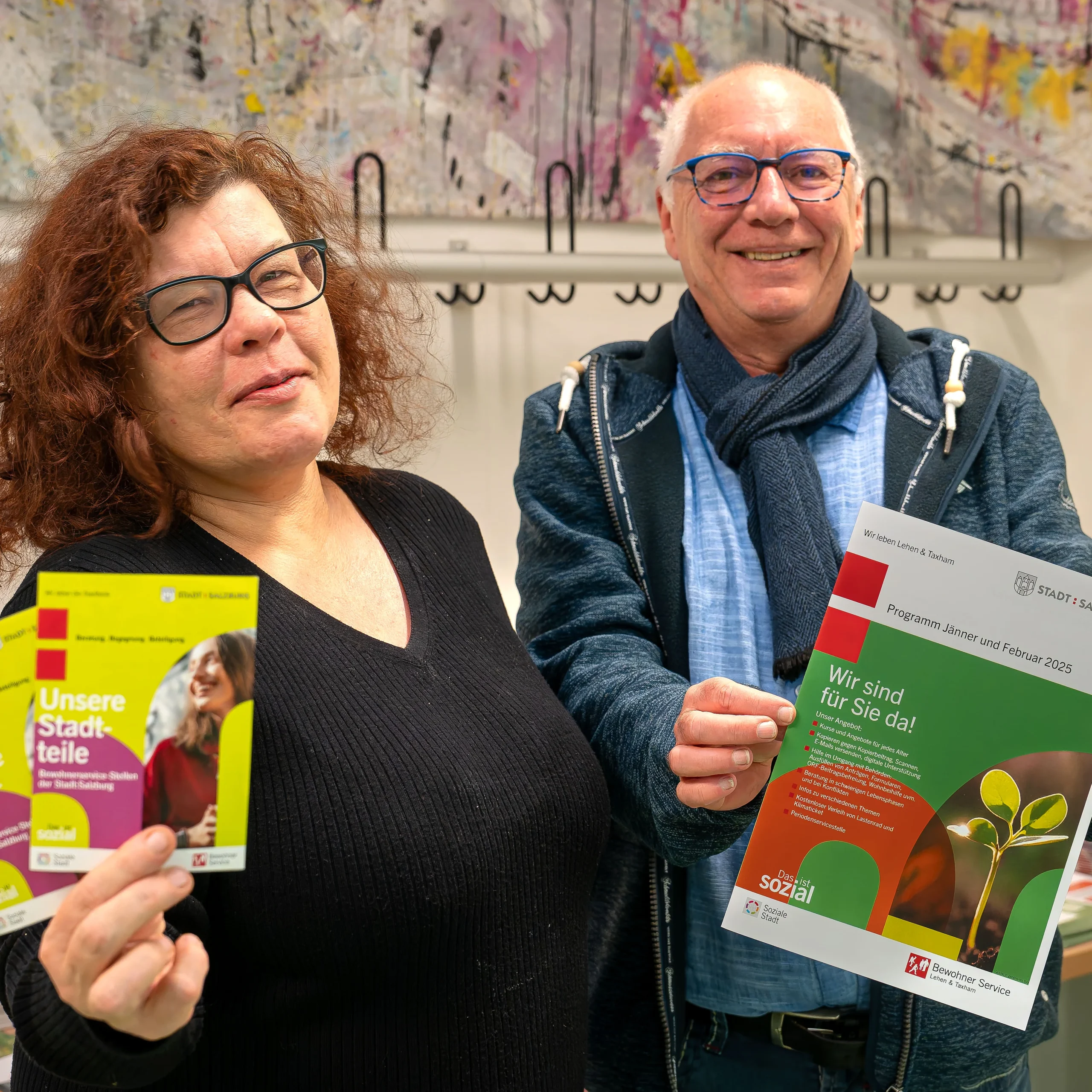 eine Frau und ein Mann aus dem betreuten Wohnen halten Magazine der Stadt Salzburg in die Kamera und lachen dabei