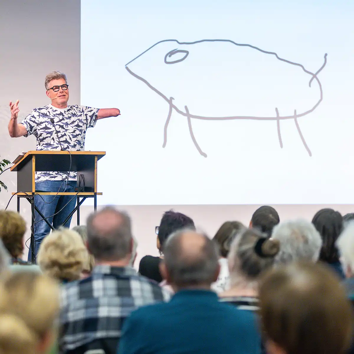 Ein mittelalter Mann mit nur einem Arm und Brille hält einen Vortrag vor Publikum und zeigt eine Zeichung