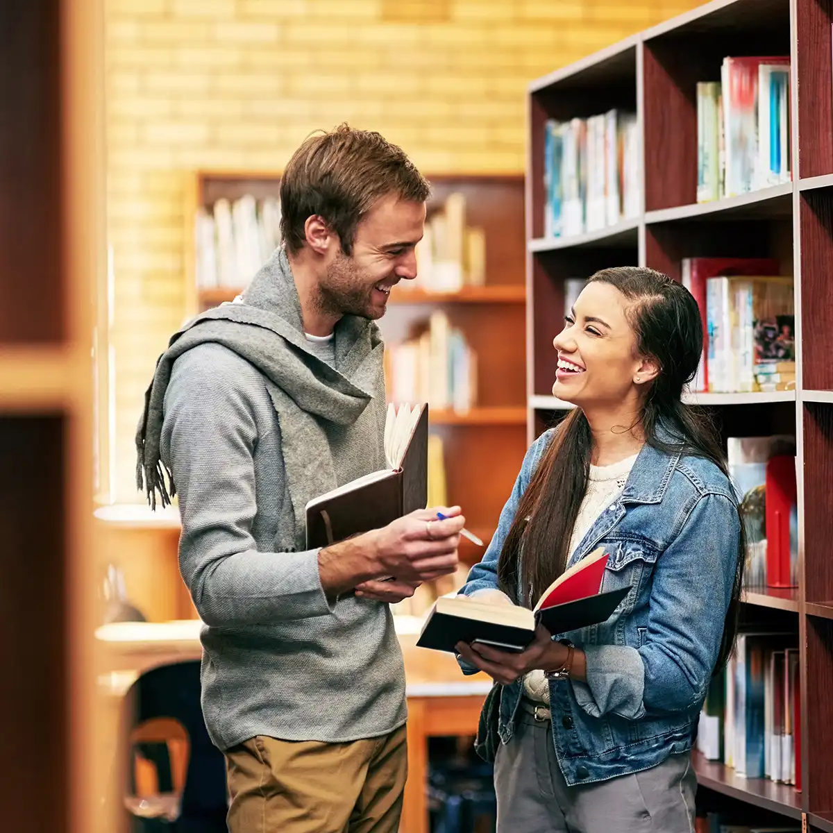 Ein Mann und eine Frau lächeln sich in einer Bibliothek an, während sie Bücher in den Händen halten